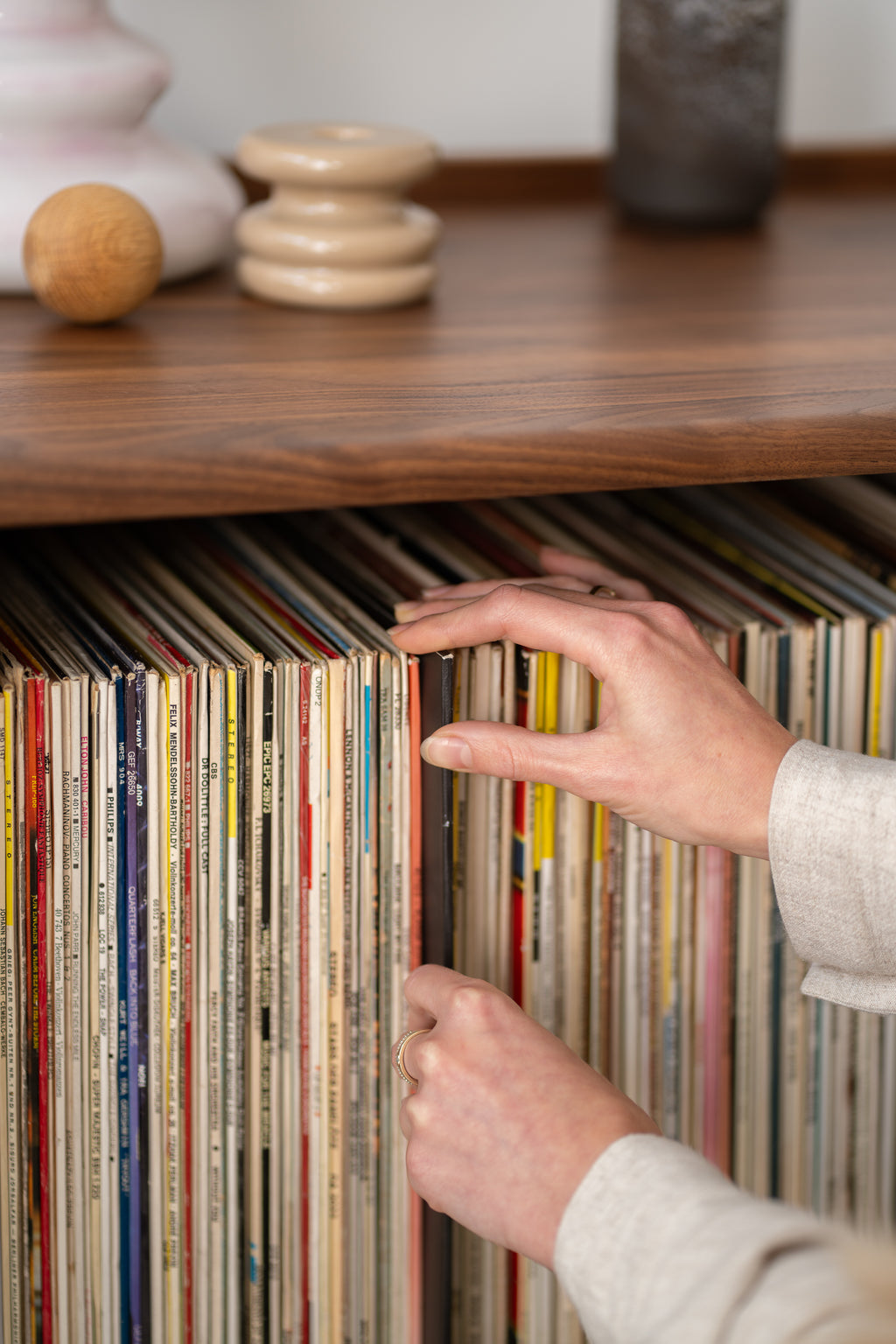 A hand browsing through a collection of vinyl records on a wooden shelf.
