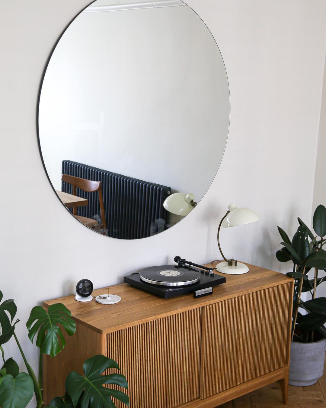 A wooden sideboard with a turntable, a lamp, and plants; on the wall, a round mirror.