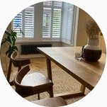 A bright interior with a wooden table and chairs, a planter with a plant on the table, and a window with blinds in the background.