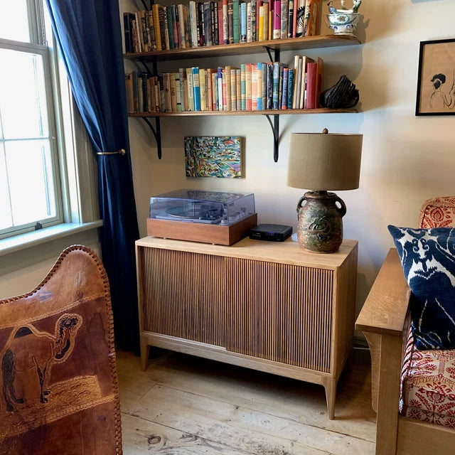 A room with a bookshelf full of books, a turntable on a sideboard, and a table lamp next to a sofa.