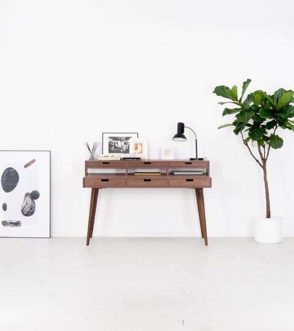 A wooden desk, a lamp, photos, and a potted plant against a white wall.