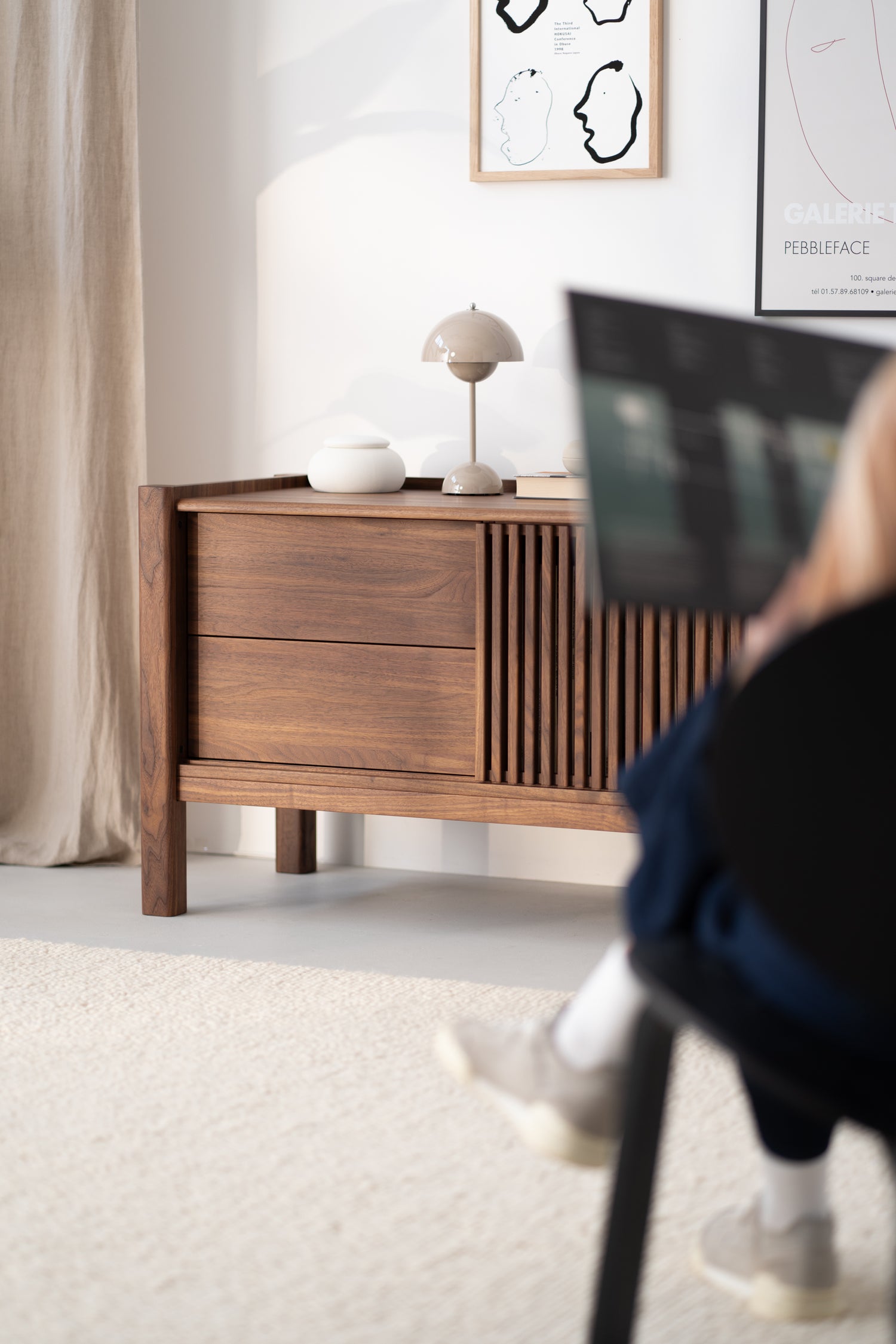 Mid-century modern wooden sideboard with decorative vases and lamp. Blurred figure using a laptop in the foreground.