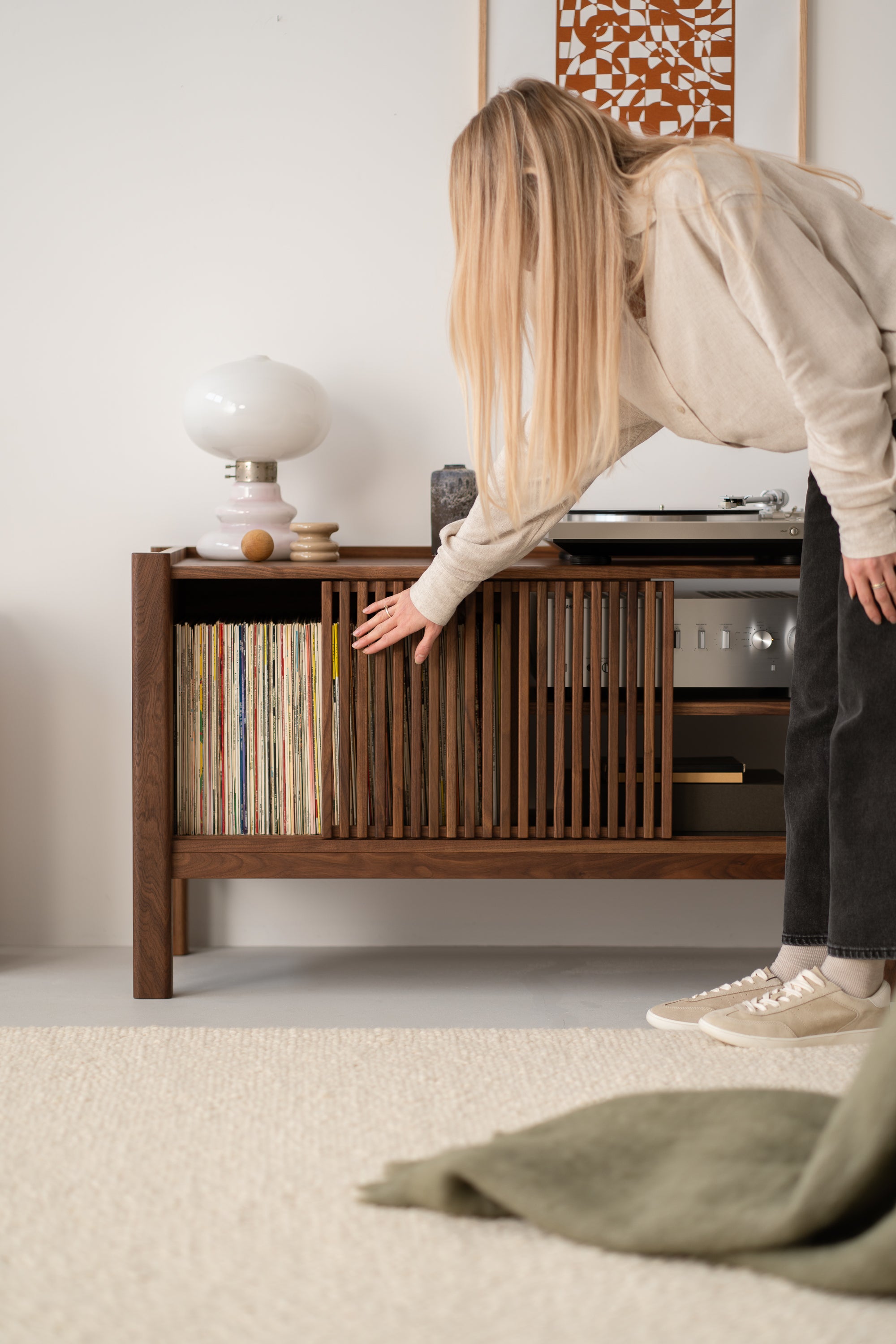 A person reaching for a book or record from a mid-century modern wooden cabinet with a lamp and decor on top.