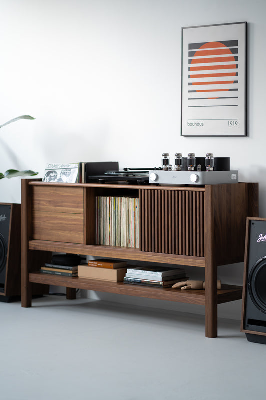 Wooden cabinet with vinyl records, a turntable, an amplifier, books, and speakers, with a Bauhaus art print on the wall.