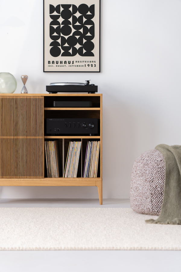Wooden cabinet with vinyl records, a turntable, and a Bauhaus poster above. A pouf with a green blanket is on the right.