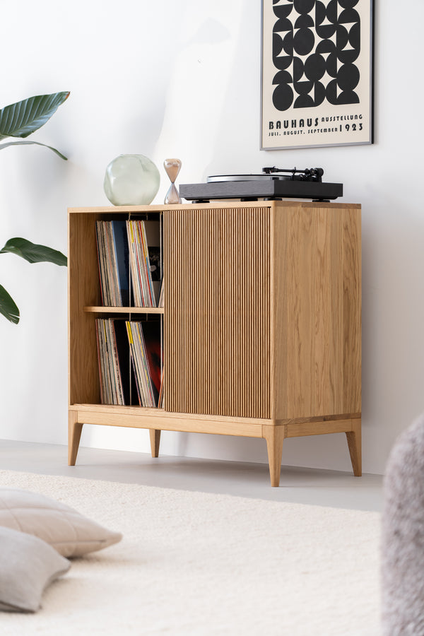 A wooden cabinet with vinyl records, a turntable, a glass vase, and an hourglass, next to a plant and a minimalist poster.