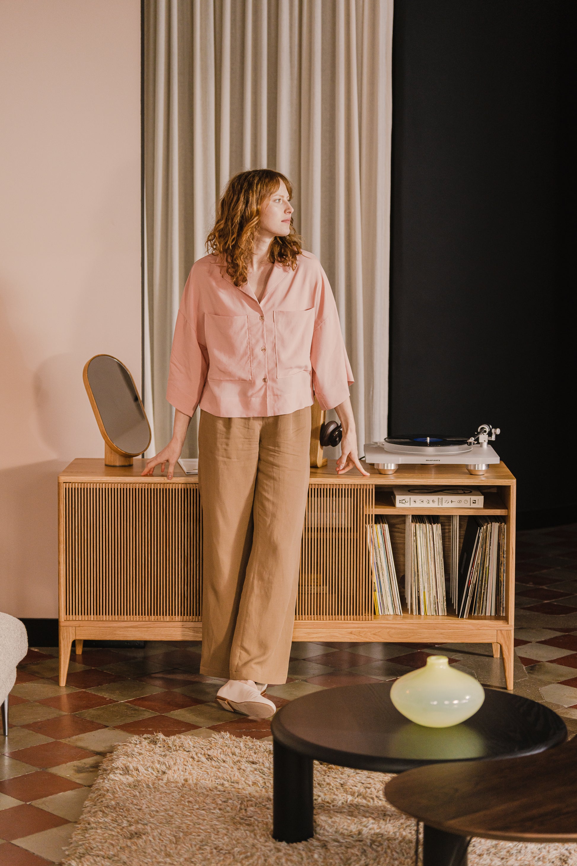 Woman in pink shirt stands by a mid-century console with a turntable, looking to the side.