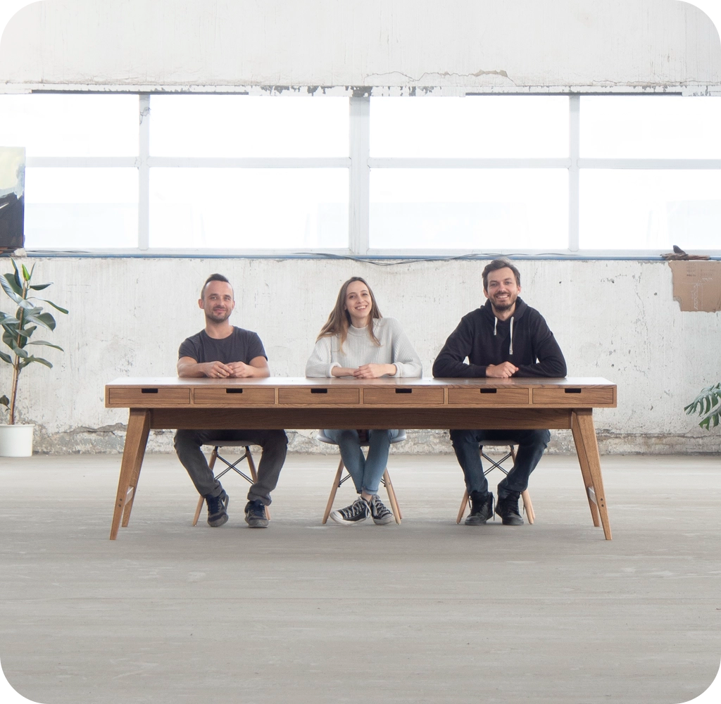Three people sitting at a large wooden table against a light-colored wall.