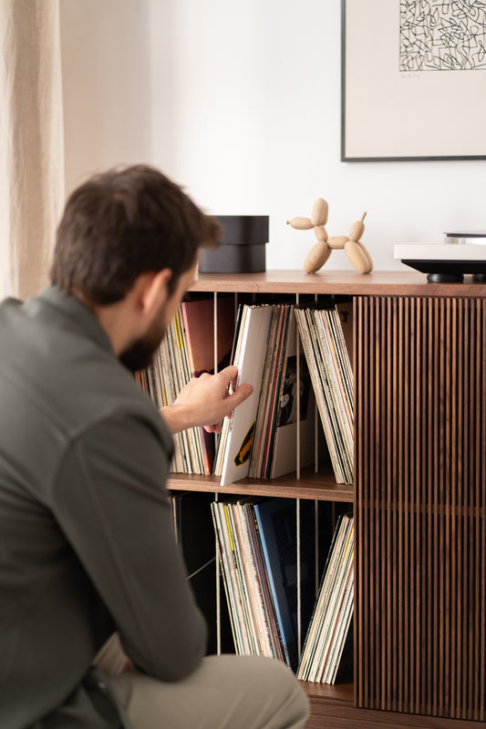 A person selects a record from a wooden shelf filled with vinyl records, beside a turntable and a decorative object.