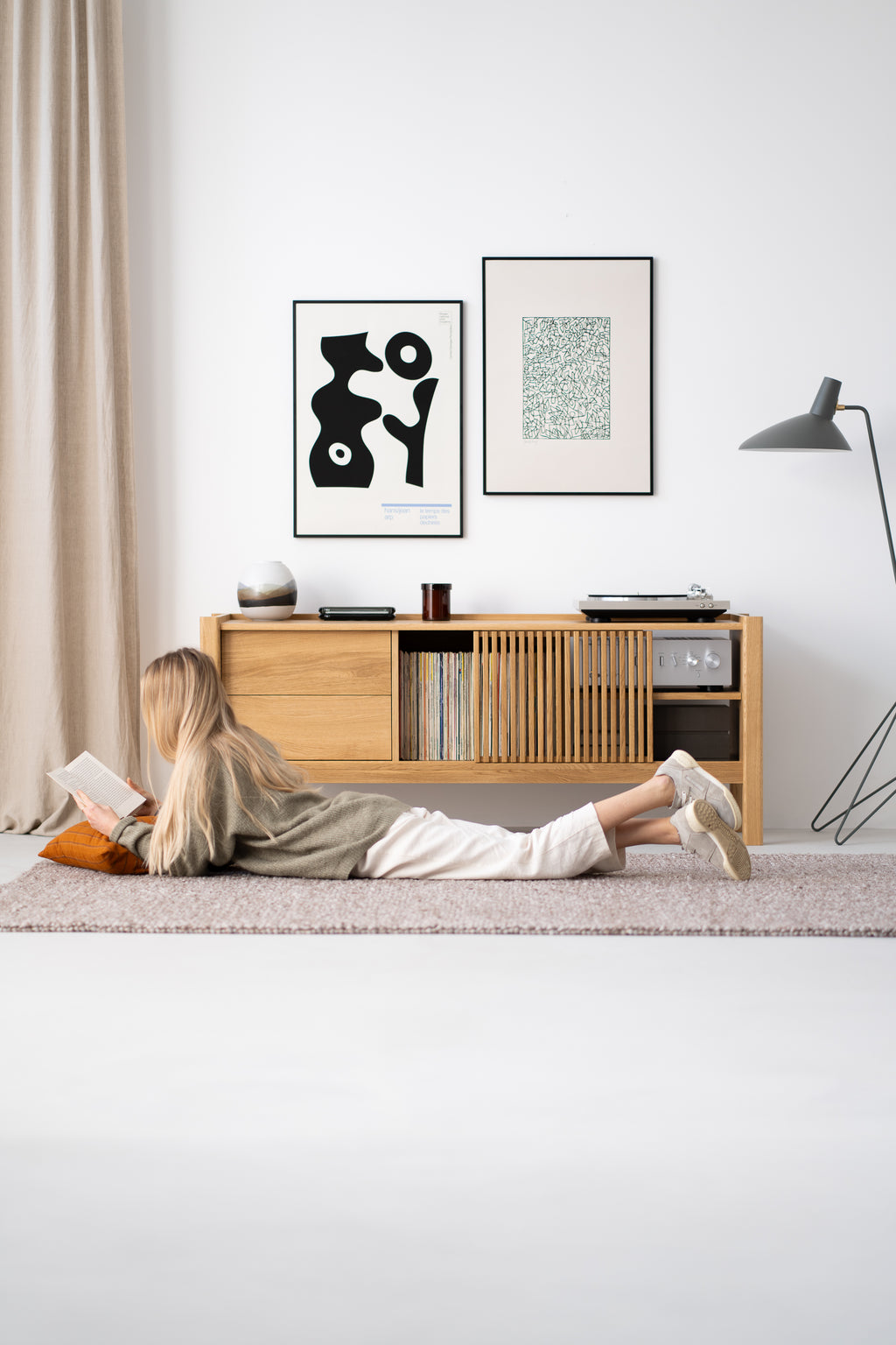A person lying on a rug reading a book, next to a wooden cabinet with paintings on the wall and a lamp.