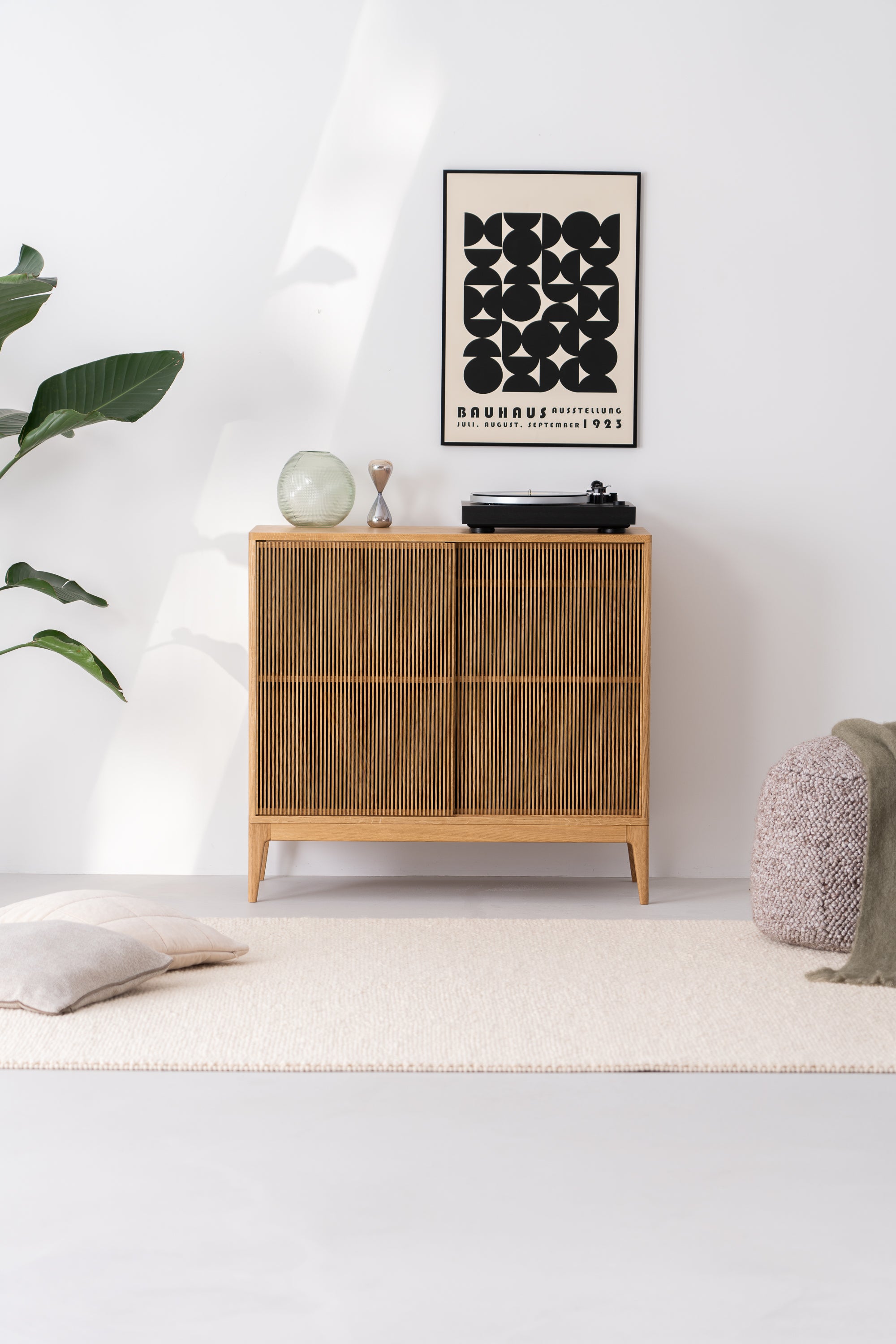 Modern minimalist room with wooden sideboard, black and white artwork, turntable, plant, cushions, and pouf.