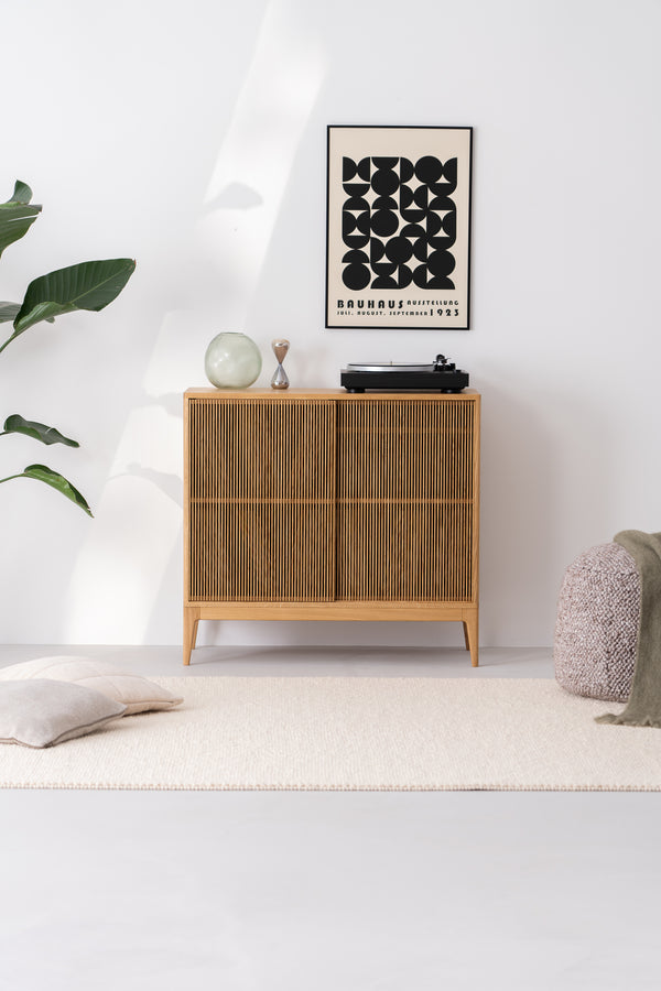 Modern minimalist room with wooden sideboard, black and white artwork, turntable, plant, cushions, and pouf.