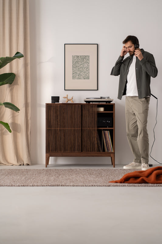 A man listens to headphones in a cozy room with a wooden cabinet, houseplant, and framed artwork on the wall.