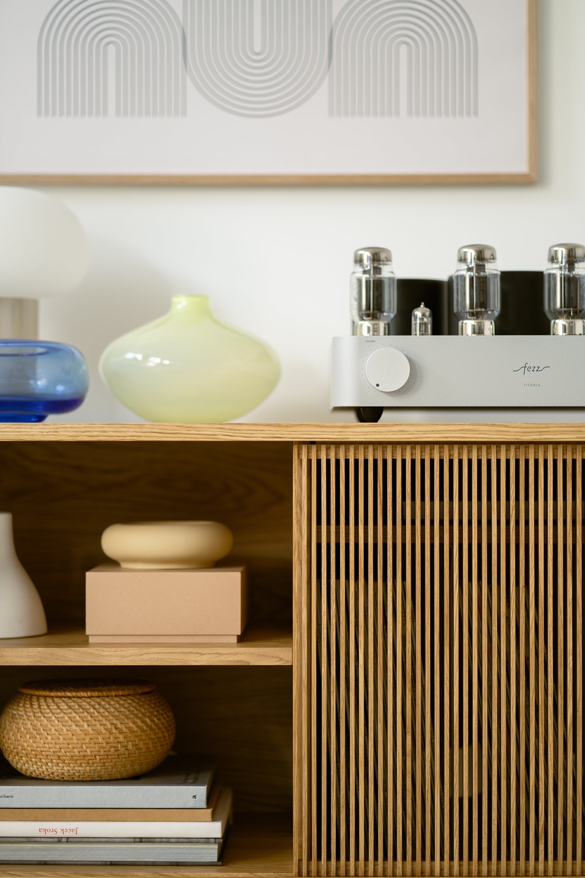 Modern wooden cabinet with vases, books, and an audio device on top, set against minimalist wall art background.