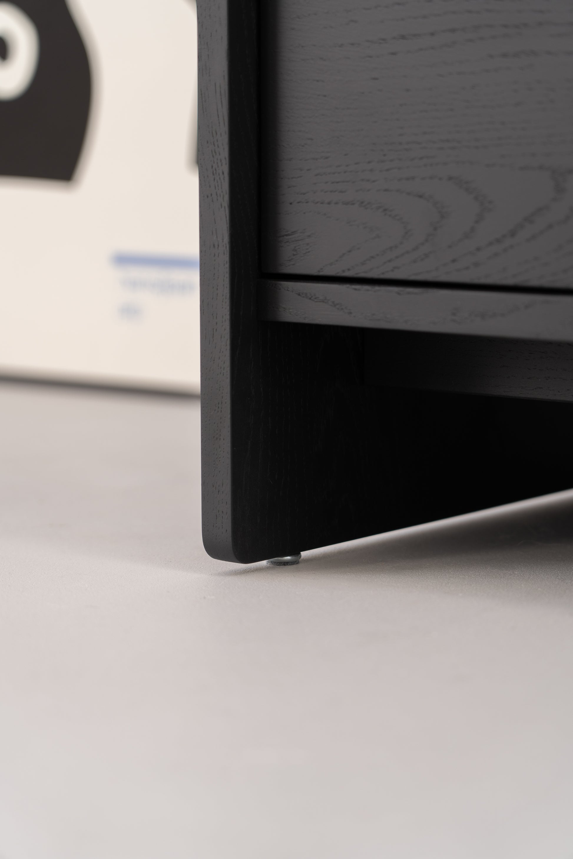 Close-up of the corner of a black wooden cabinet with adjustable metal feet on a grey floor.
