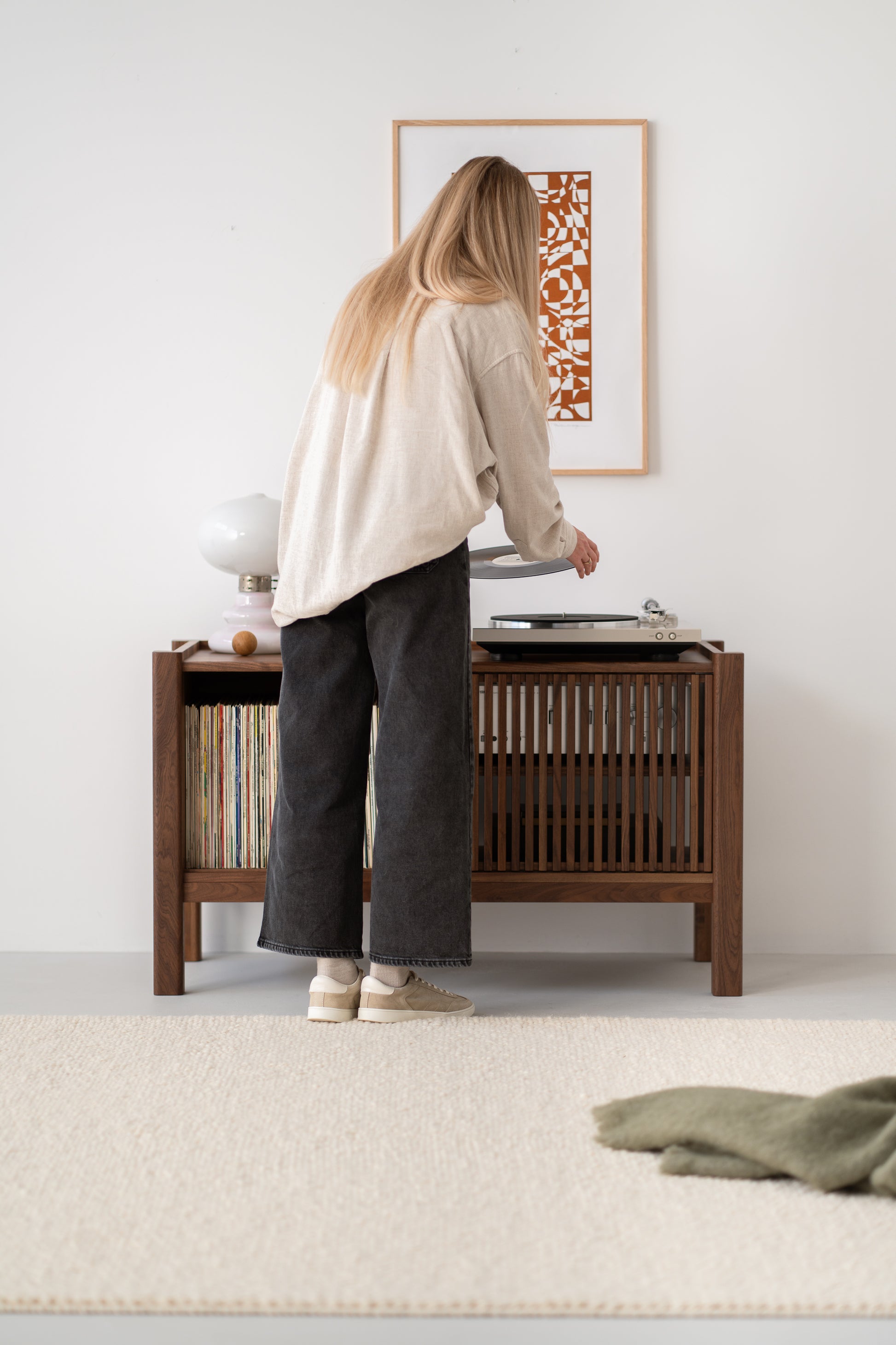 Person standing by a wooden console playing a vinyl record, with a framed abstract art piece on the wall above.