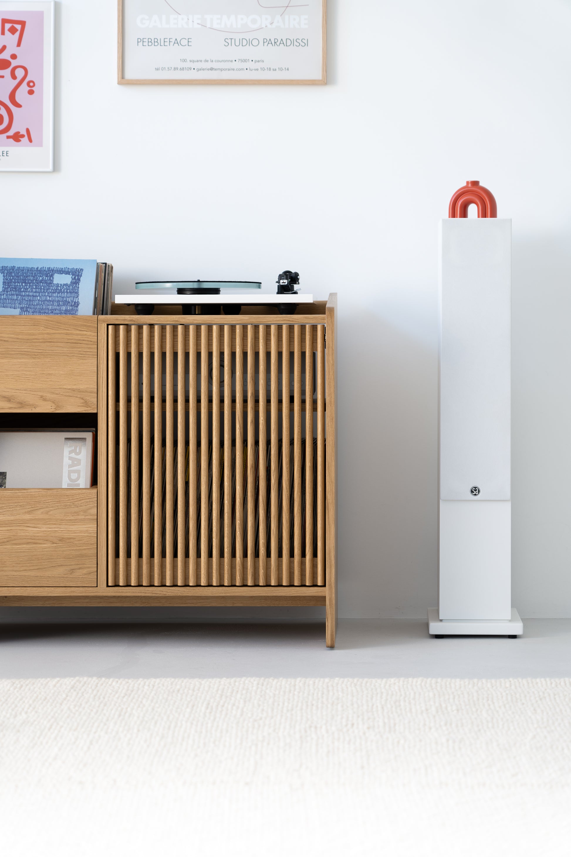 Minimalist room with a wooden cabinet, a turntable, vinyl records, framed art, a white speaker, and a red vase.