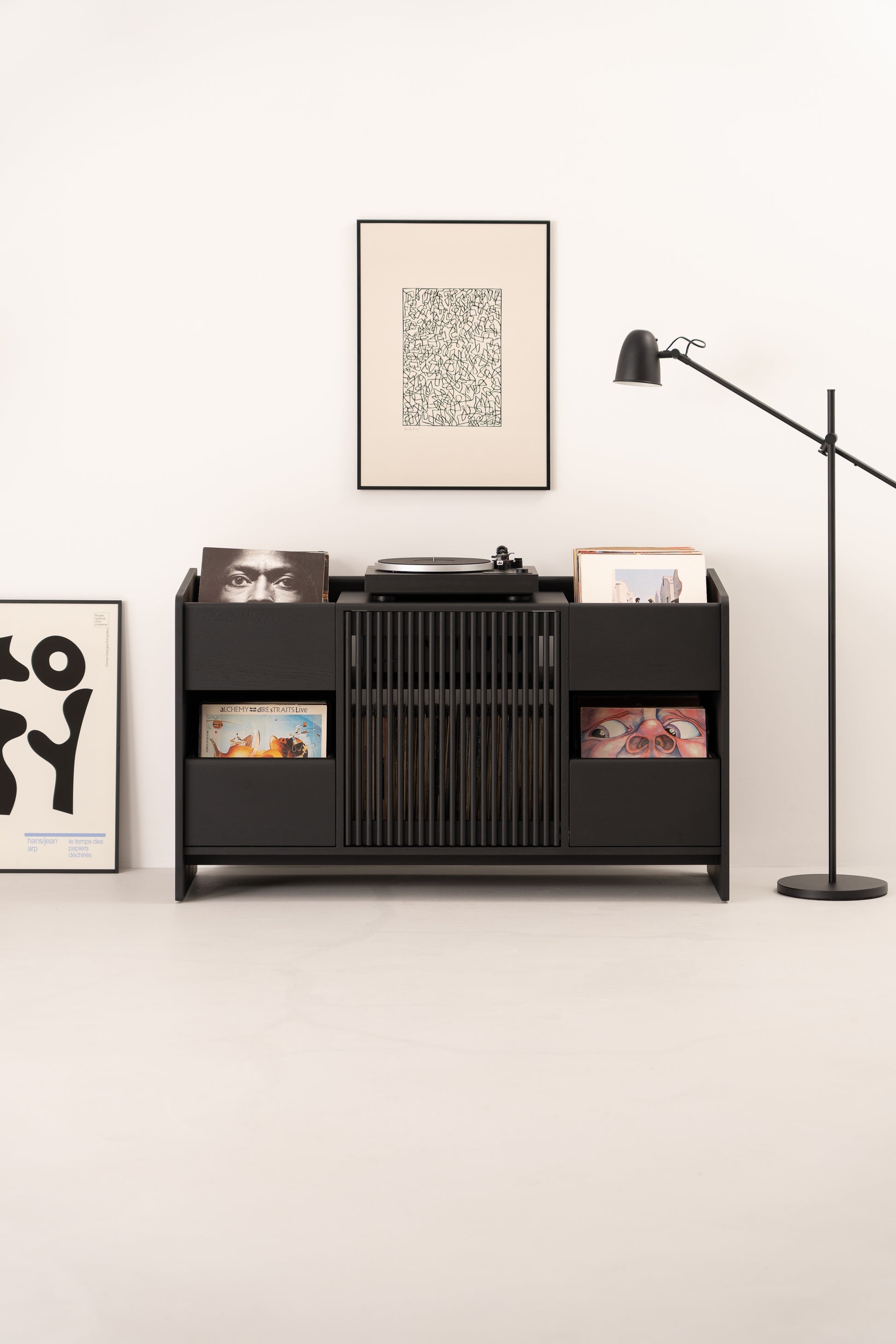 A black vinyl record cabinet with records, a turntable, a framed art piece above, and a floor lamp on the right.