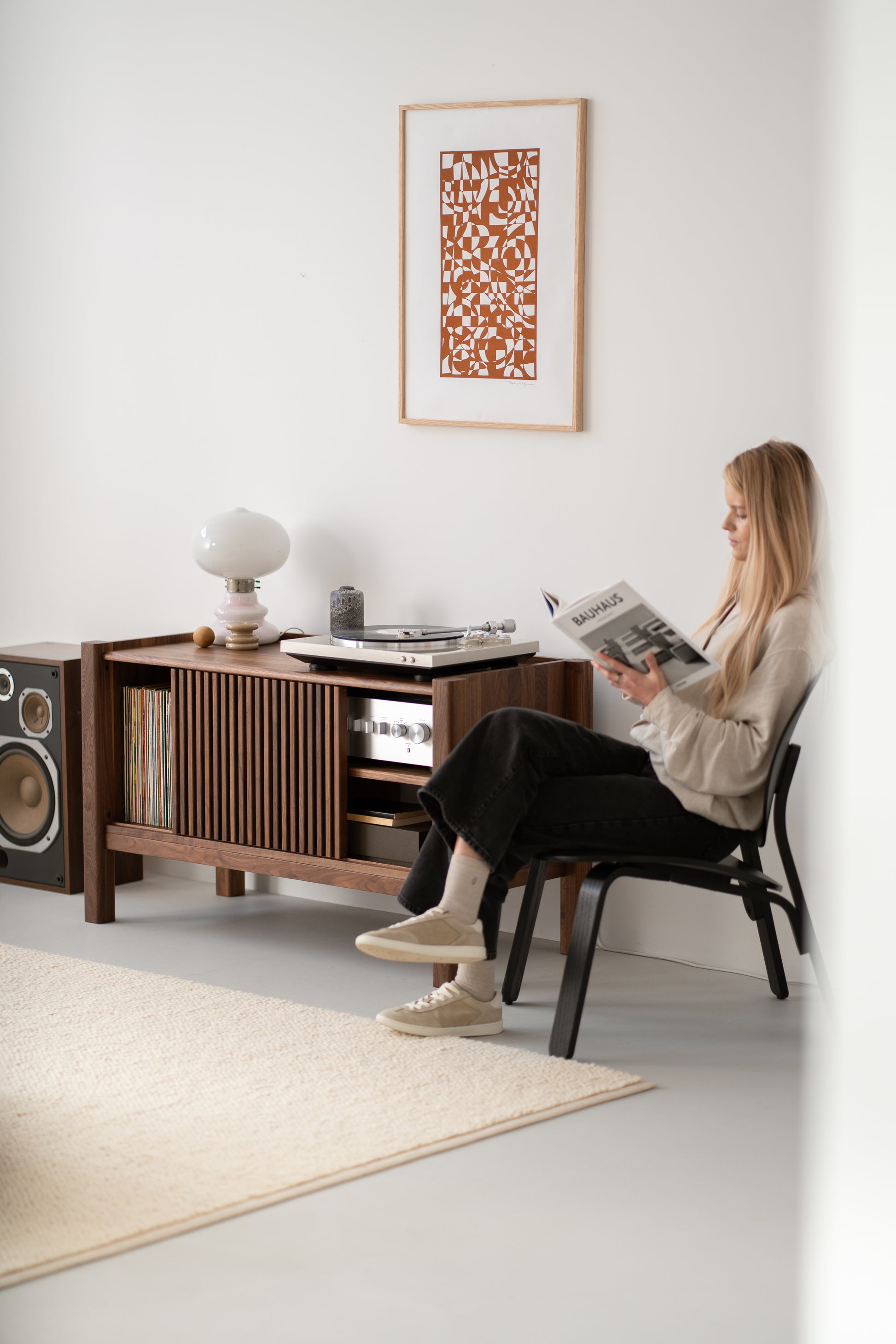 Woman reading a magazine in a modern room with a console unit, vinyl records, speakers, and abstract wall art.