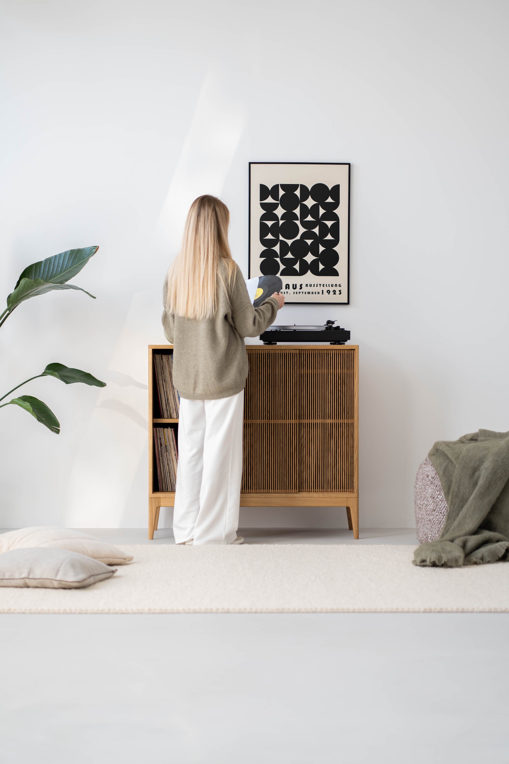 A person playing a vinyl record on a turntable next to a modern wooden cabinet, in a minimalist room with a plant.