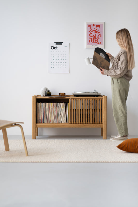 A person holding a vinyl record next to a wooden cabinet with records, a turntable, and a wall calendar showing October 2023.