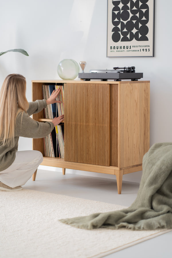 A person browsing vinyl records in a wooden cabinet with a turntable on top, next to a modern art poster.