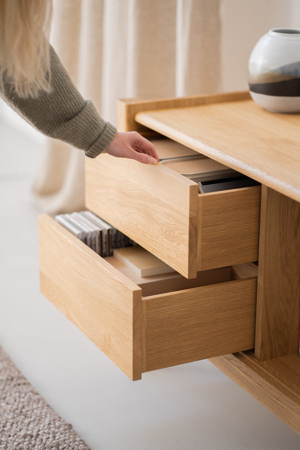 A person opening wooden drawers of a light oak cabinet, revealing books and CDs inside.