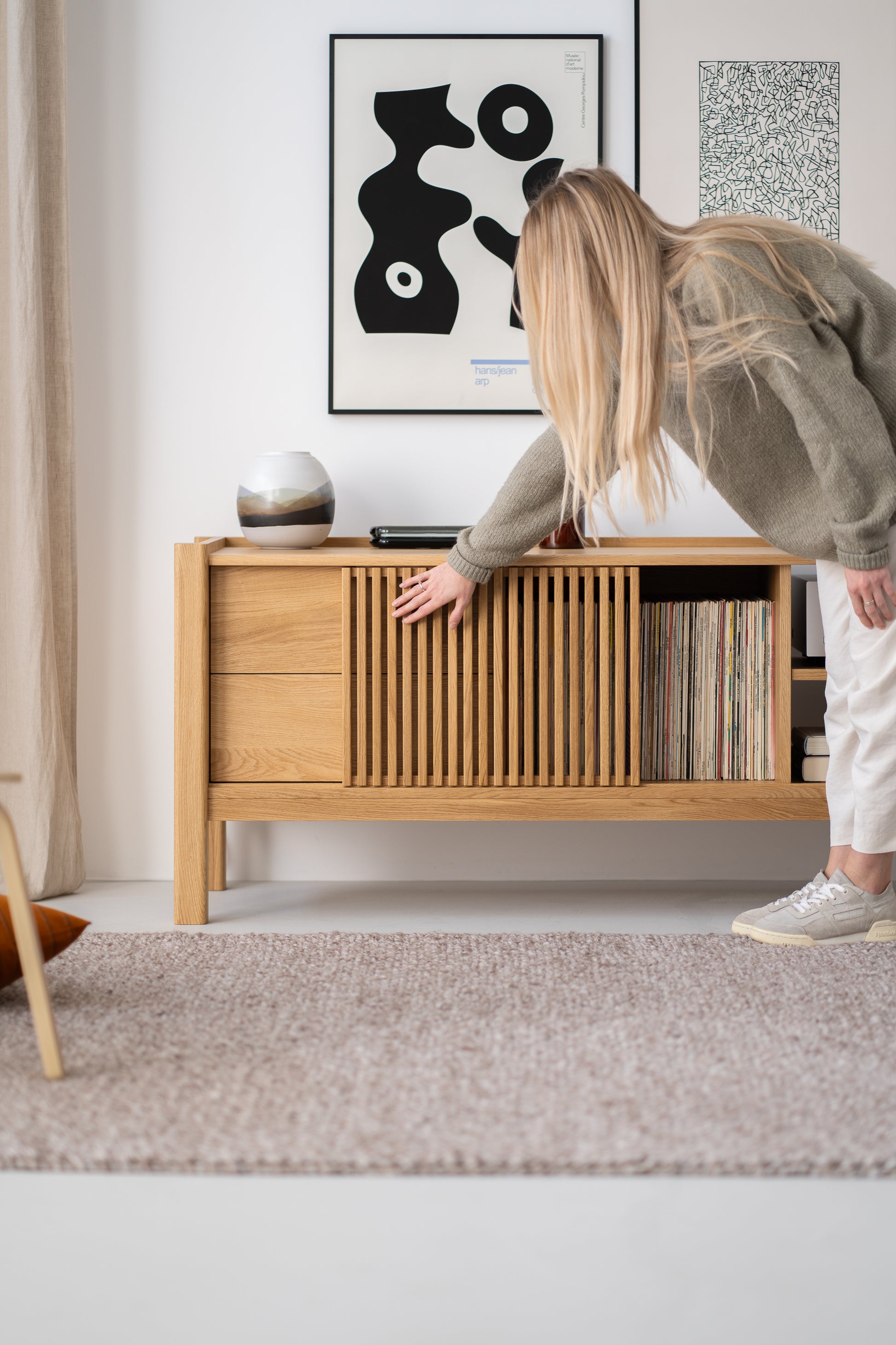 Person leaning towards a wooden sideboard, with abstract art on the wall and books or records inside the sideboard.