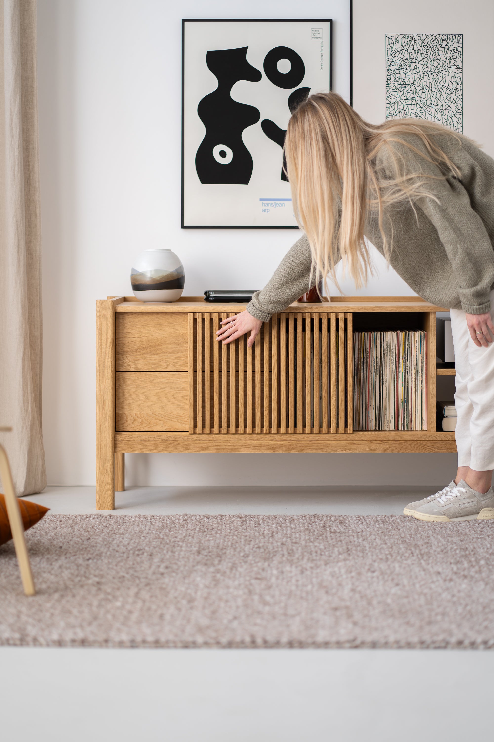 Person leaning towards a wooden sideboard, with abstract art on the wall and books or records inside the sideboard.