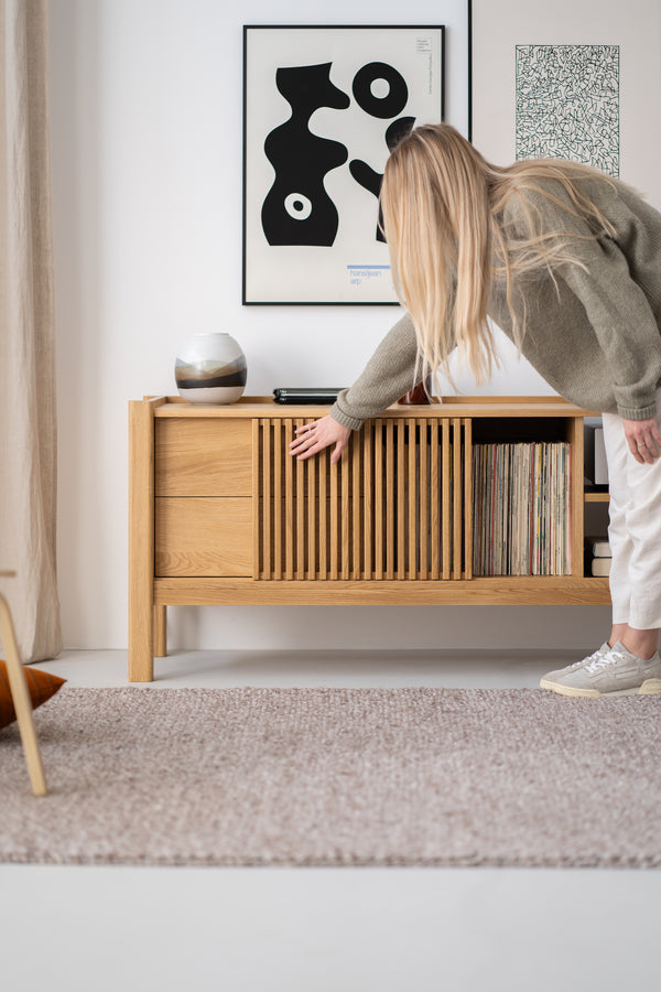 Person leaning towards a wooden sideboard, with abstract art on the wall and books or records inside the sideboard.