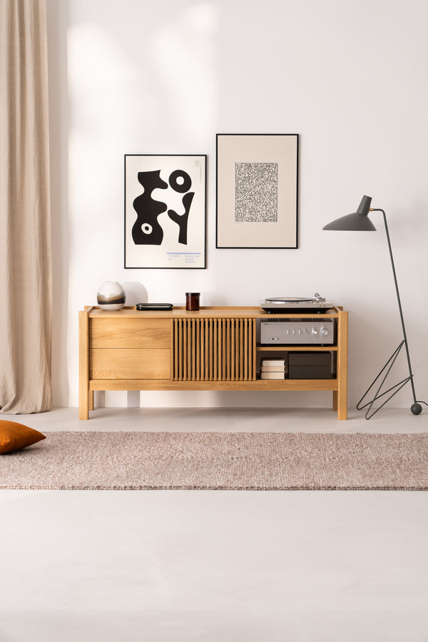 Wooden sideboard with art frames, a lamp, and decor items, set against a white wall with beige curtains and a rug.