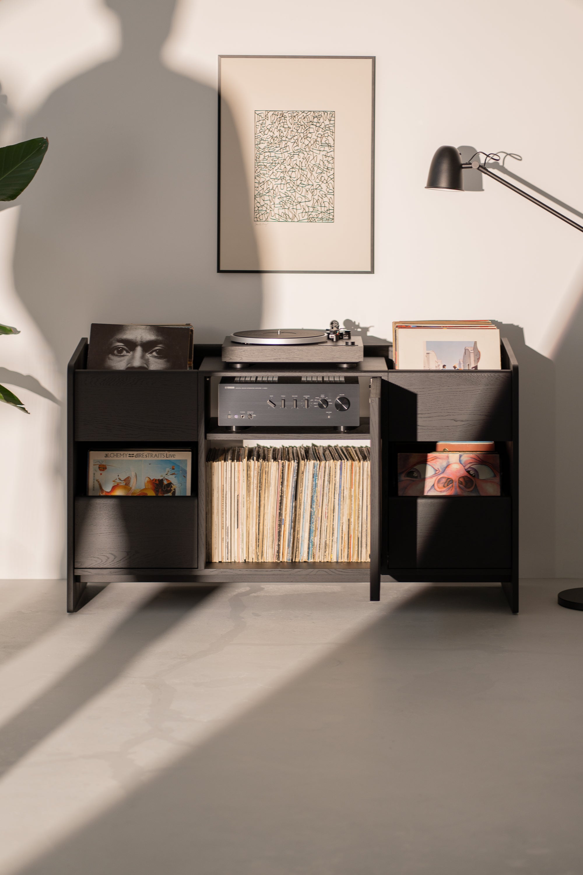A vintage record player and vinyl collection on a wooden stand, with a framed artwork and a modern lamp above.