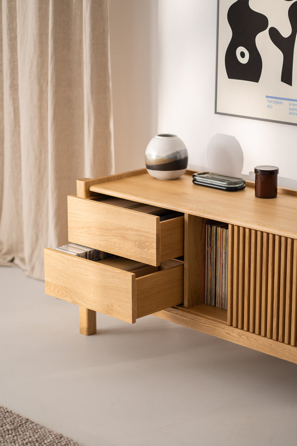 Wooden sideboard with two open drawers, ceramic vase, glass tray, and candle on top, and a framed abstract artwork above.