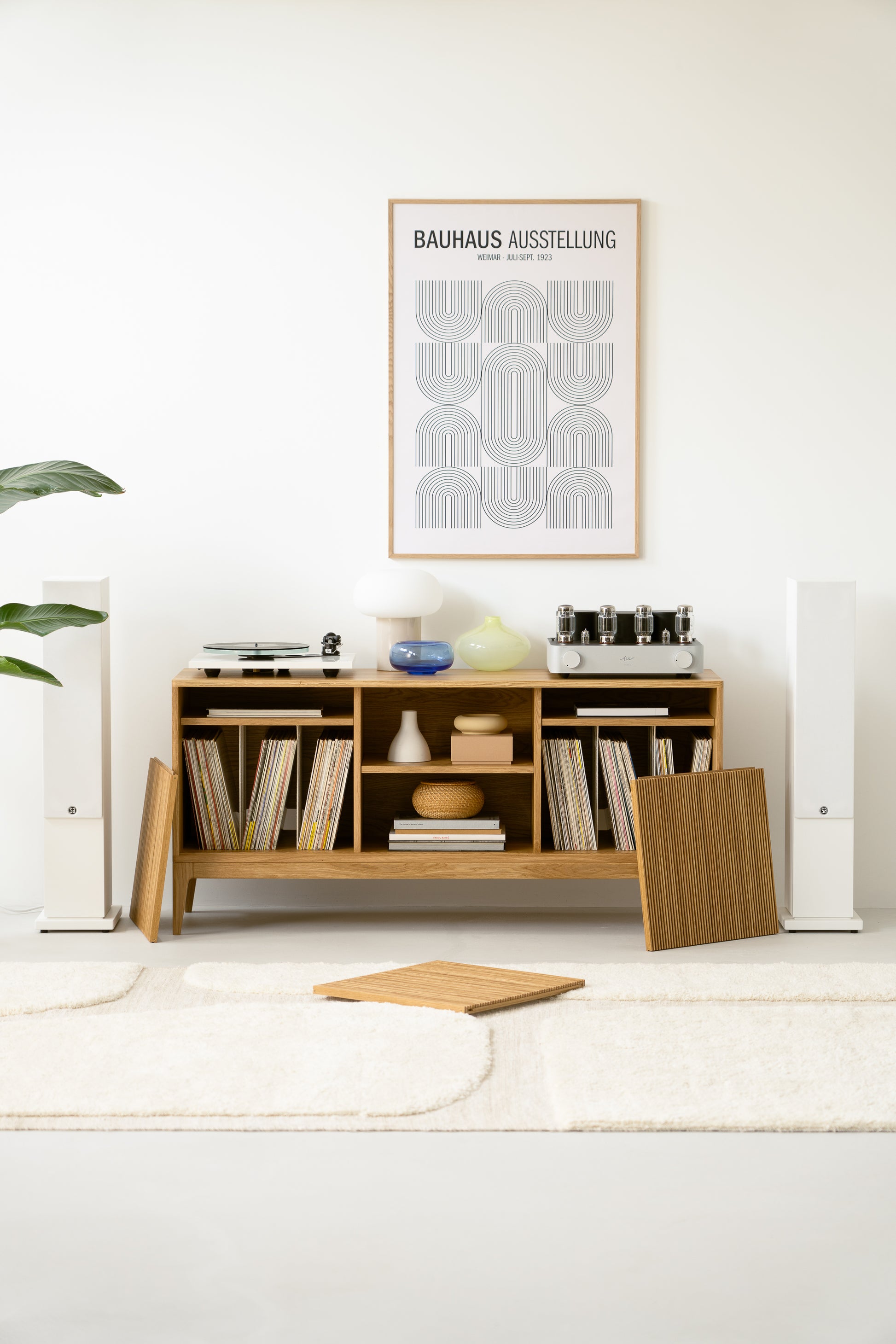 Modern living room setup with a wooden shelf holding records, decor items, and audio equipment, beneath a Bauhaus poster.