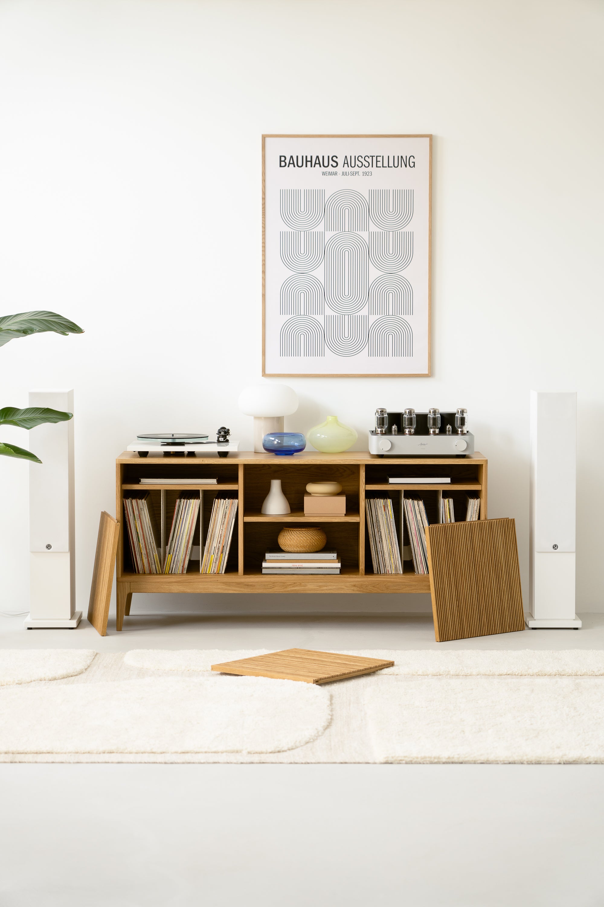 Modern living room setup with a wooden shelf holding records, decor items, and audio equipment, beneath a Bauhaus poster.