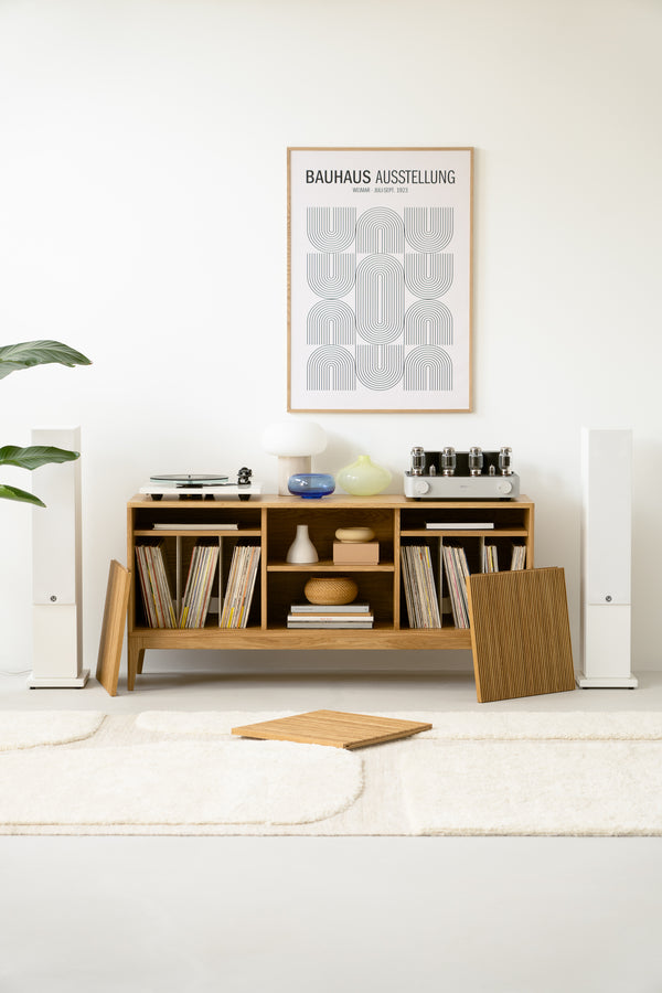 Modern living room setup with a wooden shelf holding records, decor items, and audio equipment, beneath a Bauhaus poster.