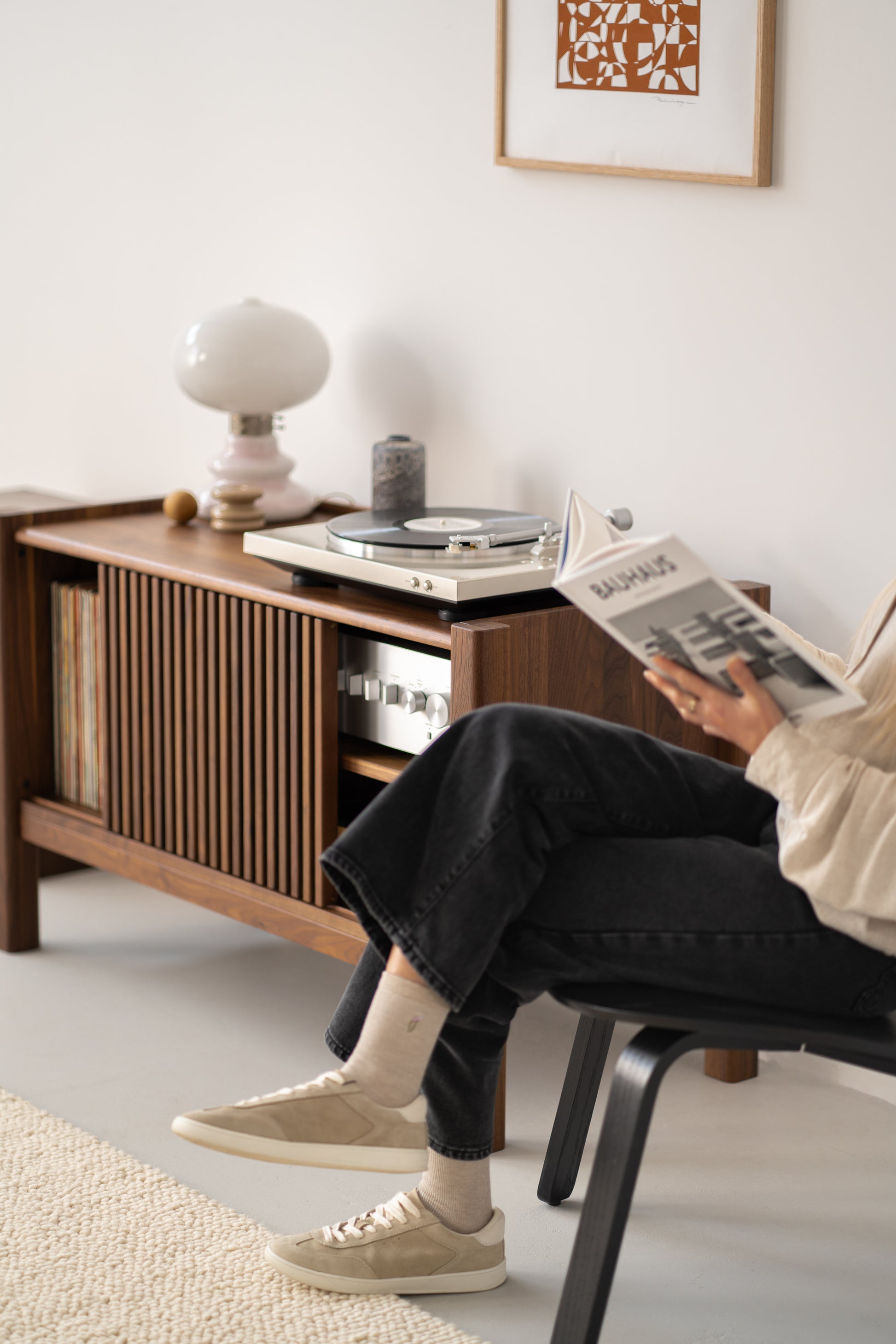Person reading a book near a wooden cabinet with a turntable and vinyl records, in a minimalist room.
