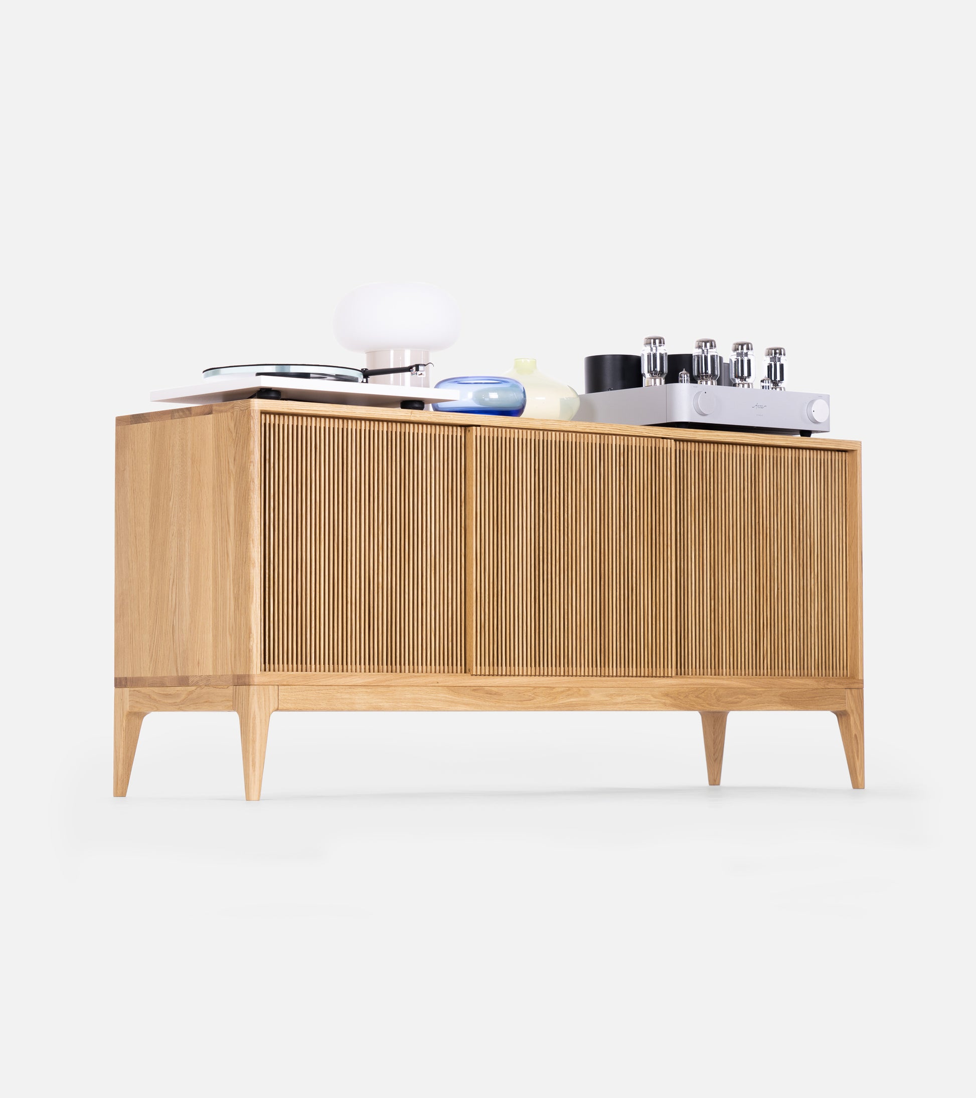 Wooden sideboard with ribbed cabinet doors, featuring a table lamp, stack of books, and various items on top.