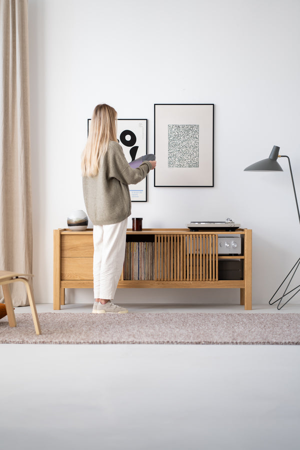 A person arranging records on a wooden console table beneath two framed artworks in a modern, minimalist room.