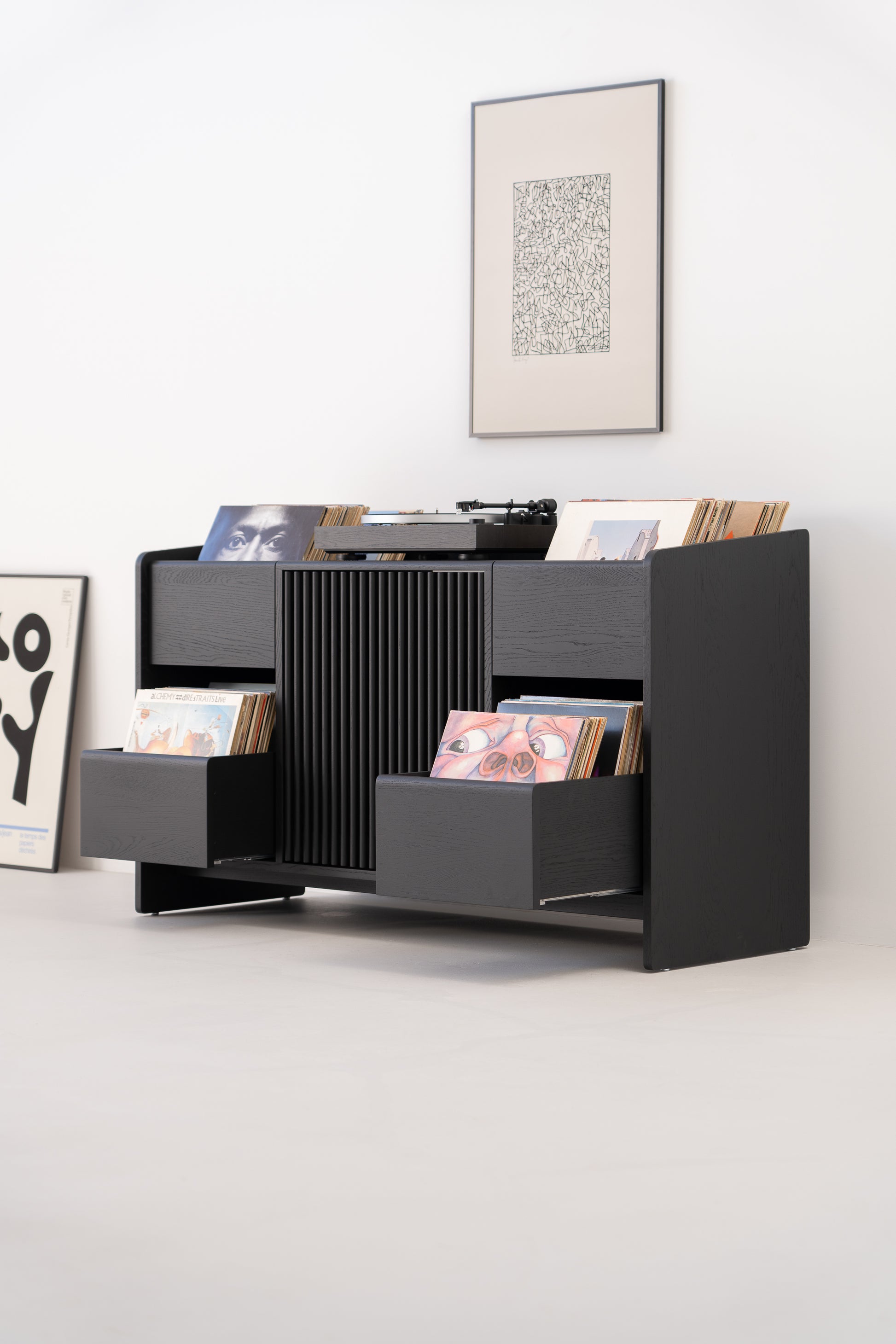 Modern black vinyl record storage unit with records, a turntable on top, and artwork on the wall behind it.