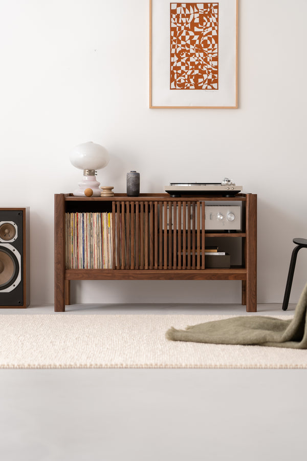 Wooden console with vinyl records, turntable, and lamp. Abstract art on wall, speaker on the left, chair on the right.