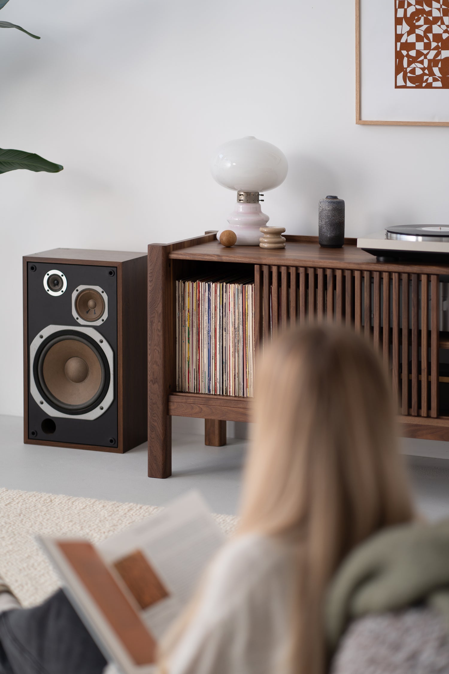 A person reading a book in a living room with a wooden shelf, a speaker, and a lamp.