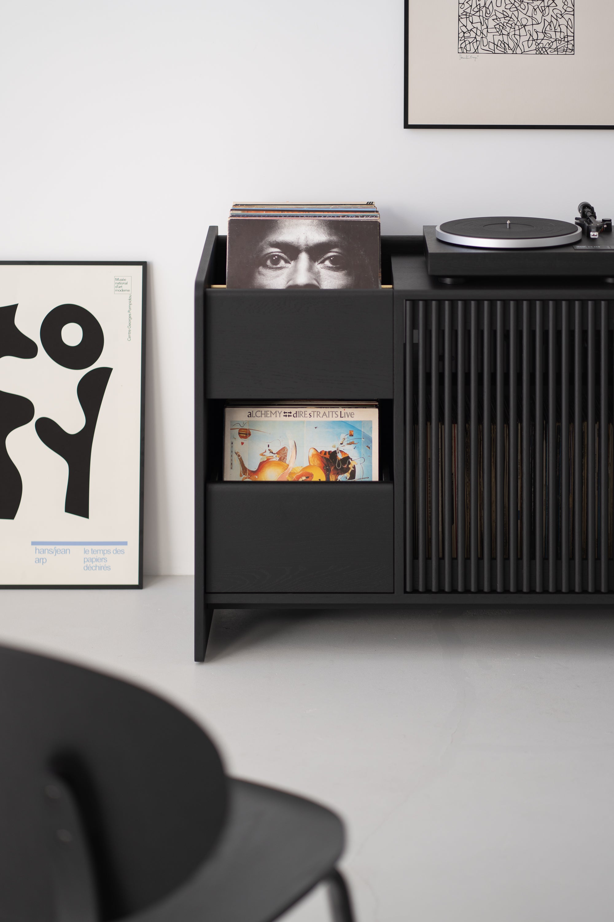 Black sideboard with vinyl records, including "Alchemy" by Dire Straits, a turntable on top, and framed art on the floor.