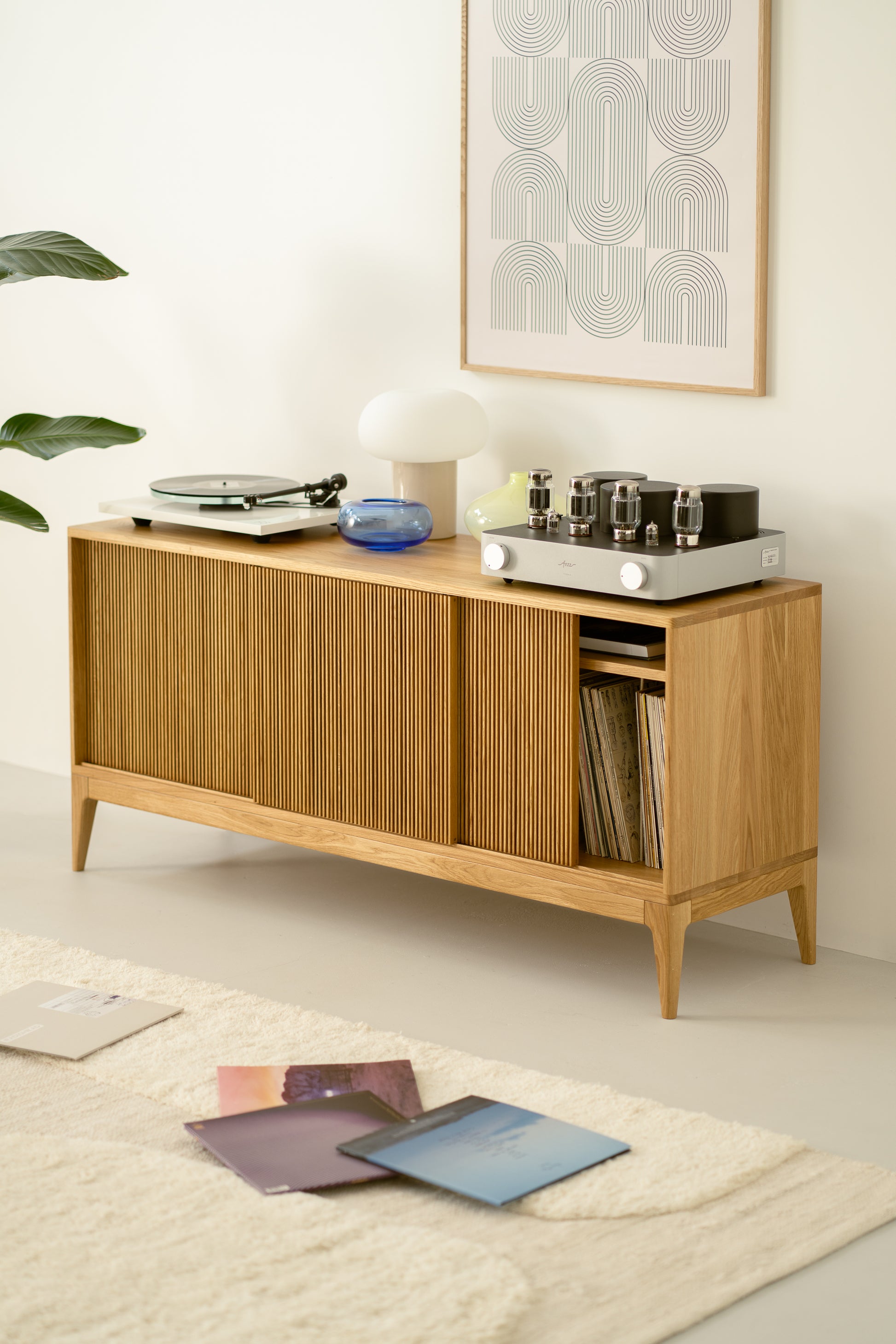 Wooden sideboard with vinyl records, a record player, and tube amplifier on top, abstract wall art, and a plant nearby.