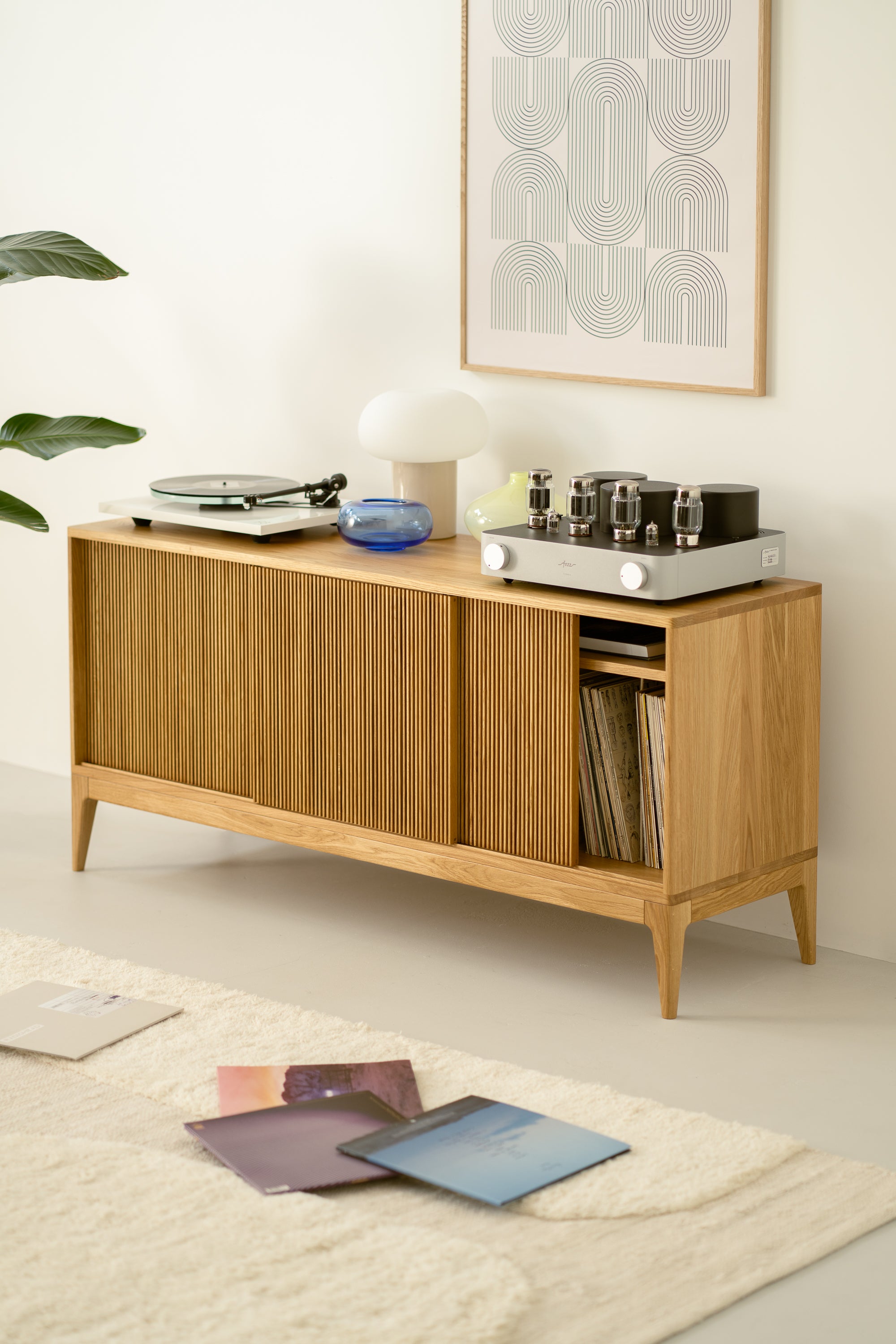 Wooden sideboard with vinyl records, a record player, and tube amplifier on top, abstract wall art, and a plant nearby.