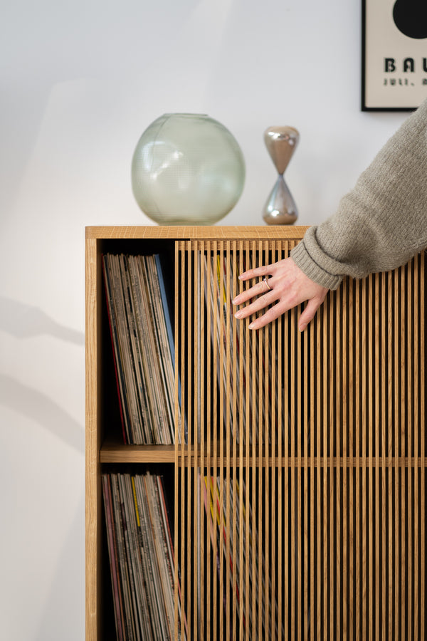 A hand slides a wooden slatted door, revealing a collection of vinyl records on a shelf.