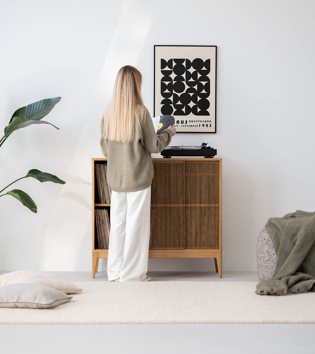 A woman standing in front of a turntable on a wooden sideboard in a minimalist interior.