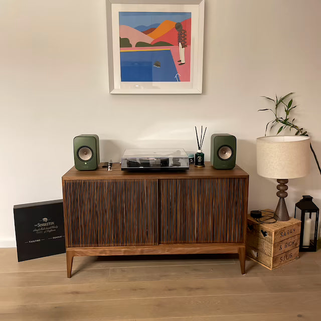 A wooden cabinet with a turntable and speakers, next to a lamp and a plant. On the wall, a colorful painting.