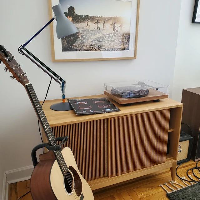 A turntable on a wooden cabinet next to a guitar and a lamp, with a painting on the wall in the background.