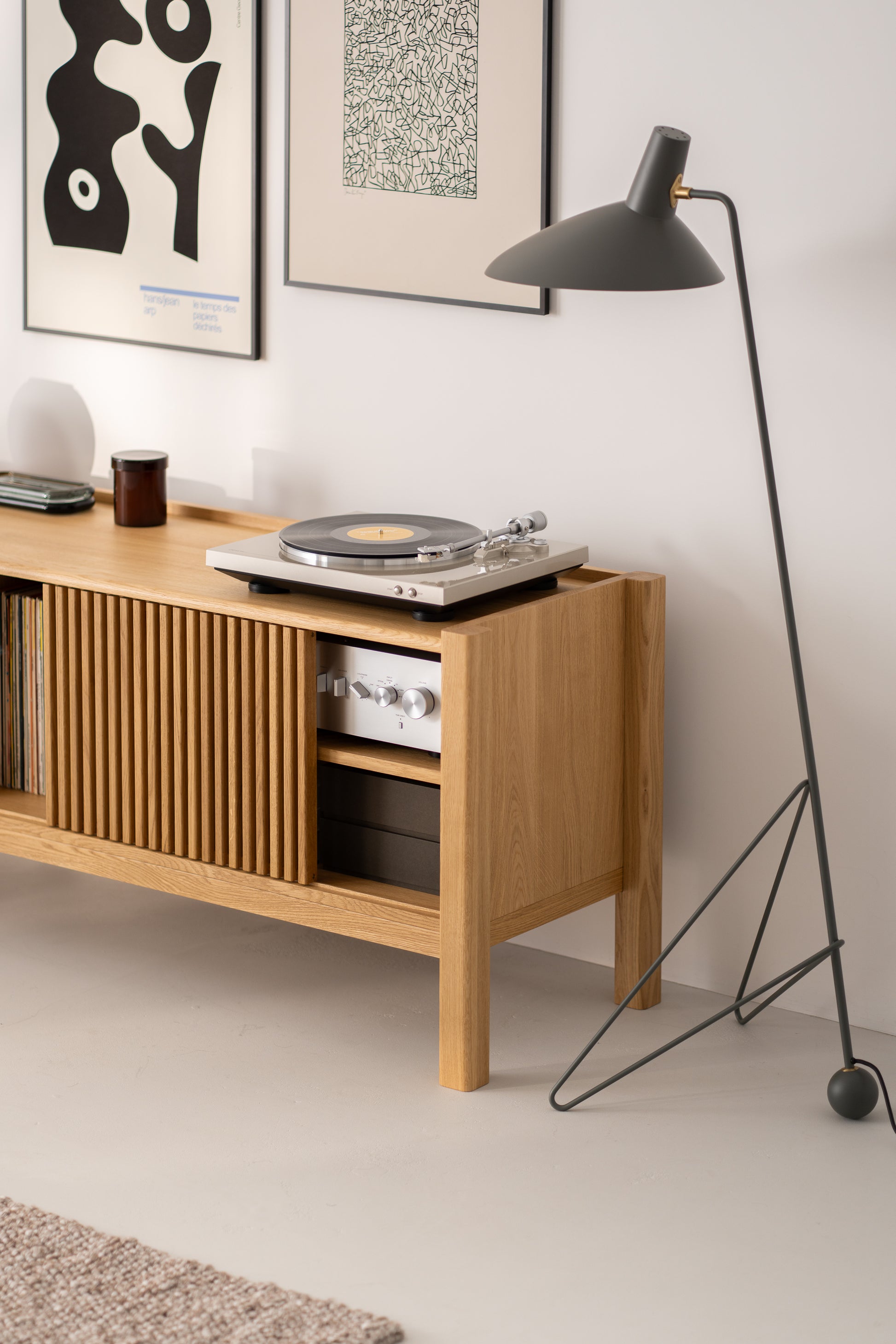 A modern vinyl record player on a wooden console table with framed abstract art above and a floor lamp beside it.